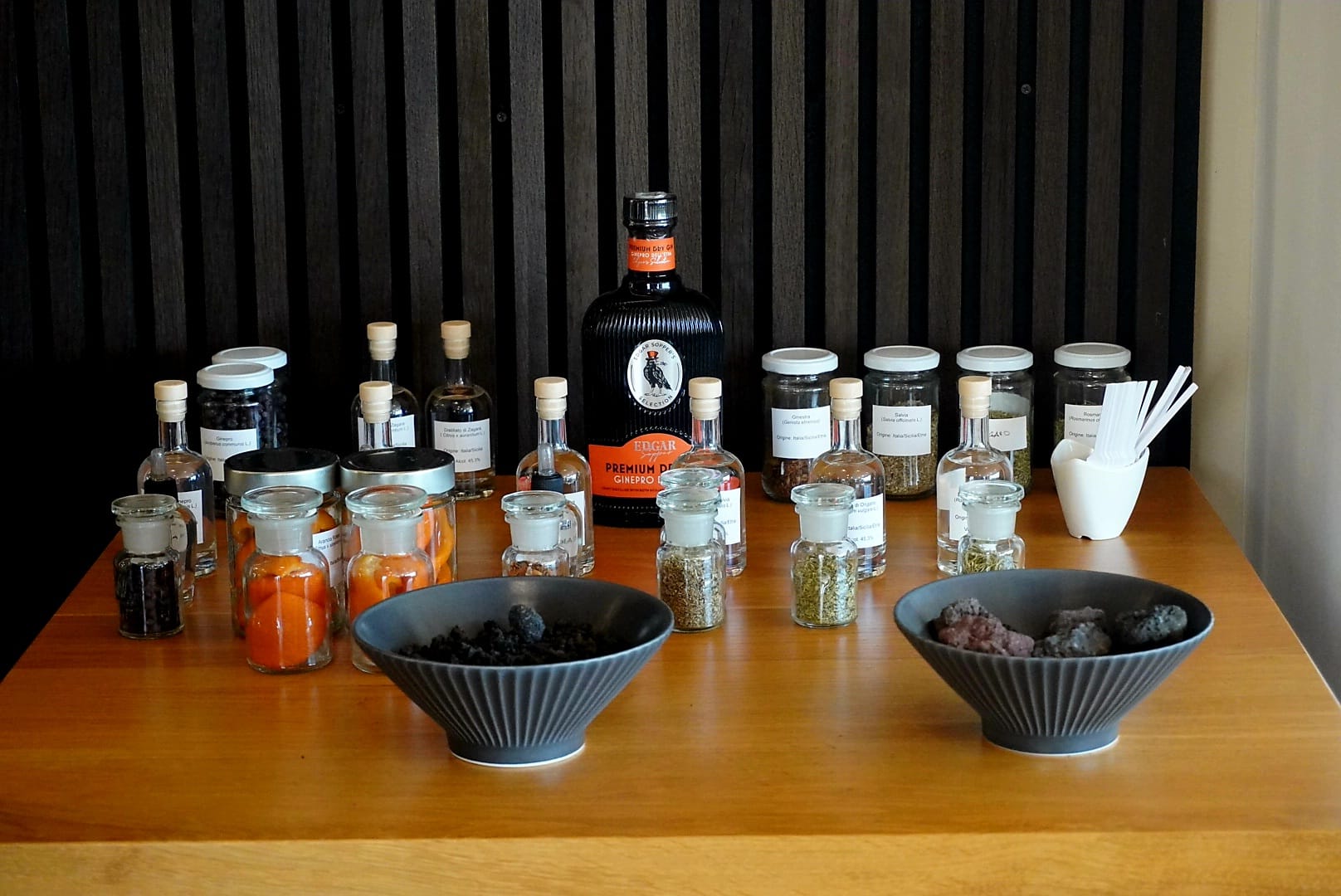 Bottiglia di Edgar Sopper Premium Gin esposta su un tavolo in legno insieme a ciotole con pietra lavica dell'Etna, arance fresche e vasetti di vetro contenenti le botaniche selvatiche del distillato.