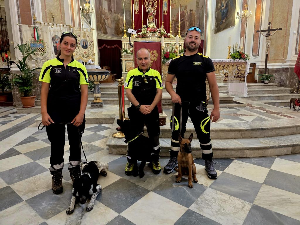 Tre volontari delle unità cinofile della Protezione Civile in divisa con i loro cani da soccorso davanti all'altare della chiesa a Mascalucia. Foto di Mari Longo.