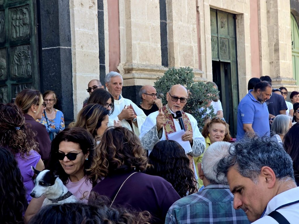Primo piano del parroco con il microfono durante la benedizione all'esterno della Chiesa di San Vito a Mascalucia, circondato dai fedeli. Foto di Mari Longo.